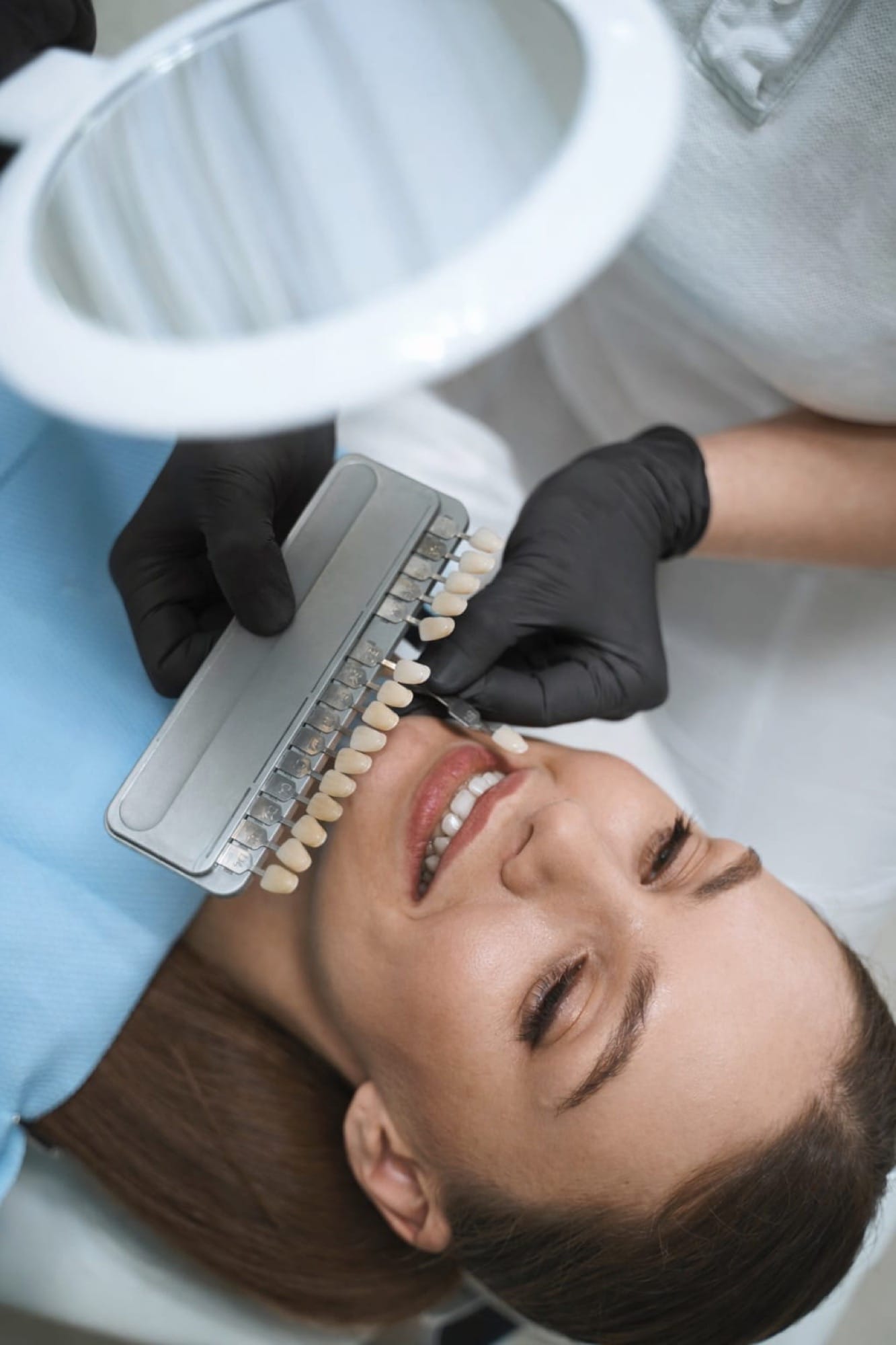 Abgleich mit Zahnfarbe: Eine Frau erhält einen Abgleich ihrer Zahnfarbe mit einem Farbfächer unter einer Lampe im Zahnarztstuhl.